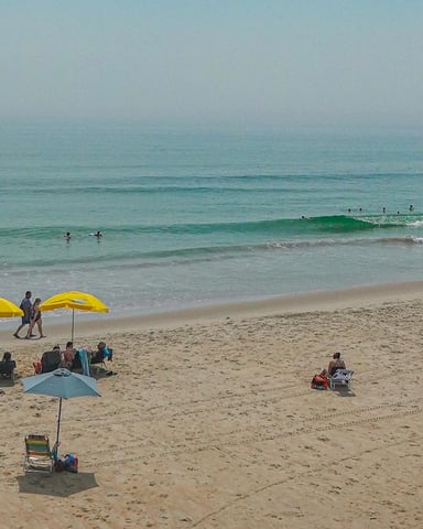 Sandy beach with swimmers and surfers in the water, beachgoers under yellow and blue umbrellas on shore