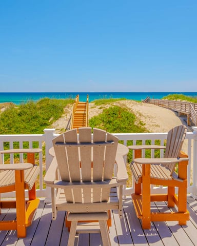 Beachfront deck with Adirondack chairs overlooking sand dunes, beach access stairs, and turquoise ocean with coastal homes nearby