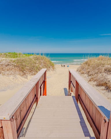 Wooden boardwalk with red railings leading through sand dunes to a wide sandy beach and turquoise ocean under clear blue sky