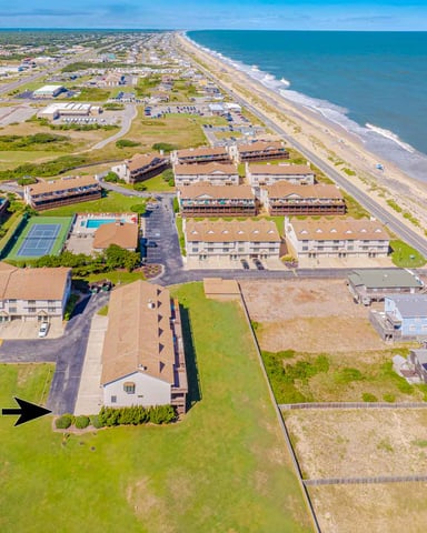 Aerial view of beachfront residential community with multi-story buildings, tennis court, pool, and direct beach access along sandy shore