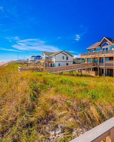 Beachfront homes on stilts with dune grass, wooden boardwalks, and sandy beach under clear blue sky