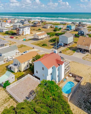 Aerial view of beachfront neighborhood with blue house with red roof and pool in foreground, sandy yards, and turquoise ocean beyond.