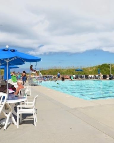 Outdoor community swimming pool with blue umbrella, lounge chairs, and swimmers in clear turquoise water
