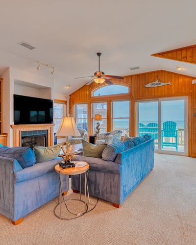 Spacious beach house living room with wood paneling, blue upholstered seating, fireplace, and ocean view through glass doors