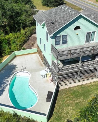 Aerial view of a two-story mint green house with a grey roof, featuring a kidney-shaped swimming pool and a small putting green, surrounded by dense greenery on one side and a road on the other.