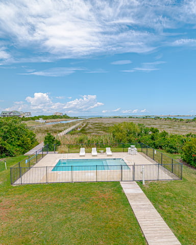 Fenced swimming pool with lounge chairs overlooking marsh, boardwalk, and waterfront homes under blue sky