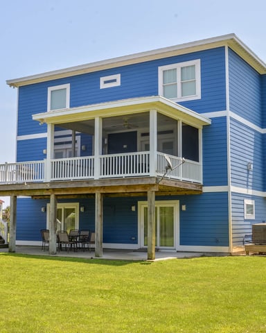 Blue two-story house with white trim, elevated on pilings, featuring a wraparound deck and ground-level porch with chairs, set on a grassy lawn.