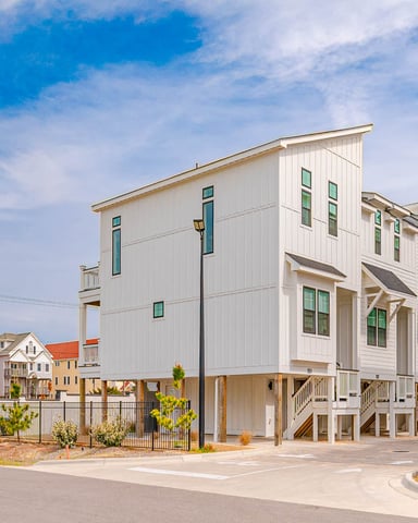 Modern white beach houses on stilts with green windows and metal railings, surrounded by colorful residential homes under blue sky.