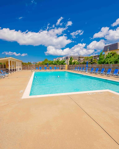 Resort pool with turquoise water, blue lounge chairs, wooden fence, and multi-story buildings under clear blue sky