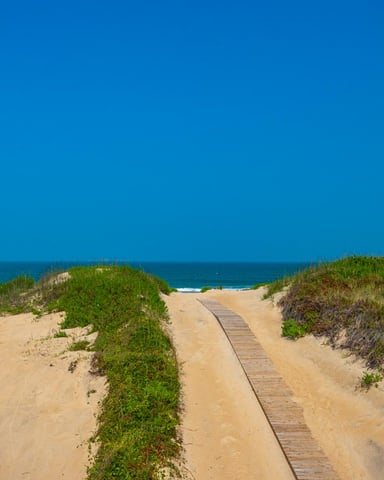 Wooden boardwalk crossing sand dunes with beach grass toward ocean, shingle cottage visible on right