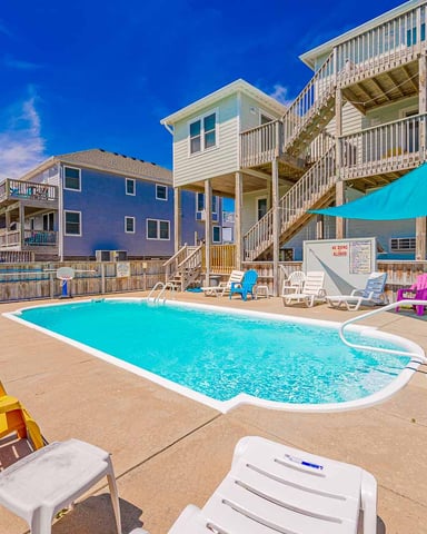 Beachfront vacation rental with swimming pool, colorful Adirondack chairs, and multi-story houses under clear blue sky