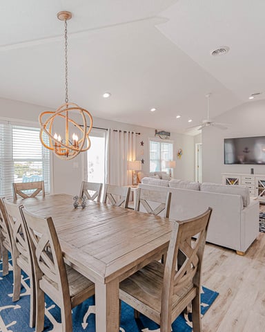Open-plan dining and living room with wooden table, orange pendant light, and ocean view through sliding glass doors.