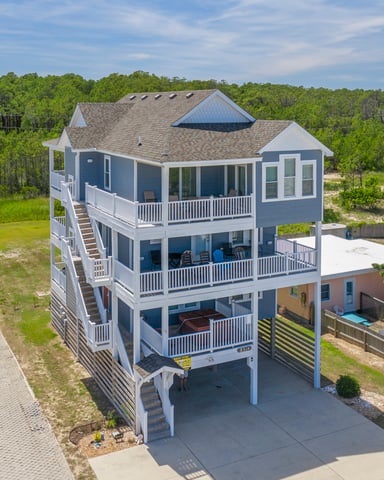 Elevated gray and white beach house with wraparound decks, carport, and ocean views, surrounded by pine trees and neighboring vacation homes.