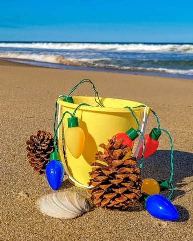 Yellow beach bucket with colorful ornaments and pinecones on sandy beach with ocean waves