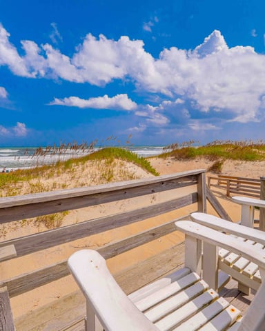 Wooden deck with white Adirondack chairs overlooking sandy beach, dunes, ocean, and beachfront house under blue sky
