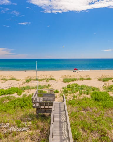 Wooden boardwalk through dune vegetation leading to sandy beach and turquoise ocean under blue sky