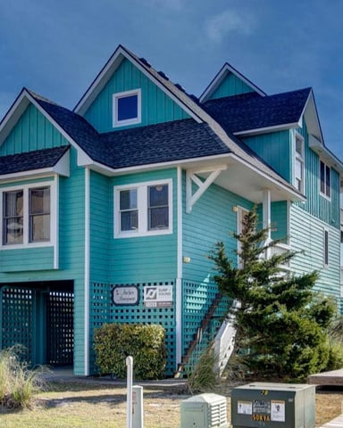 Frontal view of a two-story house painted in teal with white trims and a dark grey roof, featuring multiple windows and a staircase leading to an upper deck, situated next to a sandy pathway with shrubs on either side