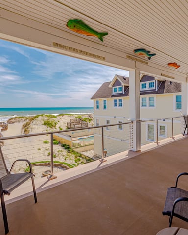 Covered oceanfront deck with lounge chairs, metal railing, and views of beach houses, sand dunes, and turquoise water