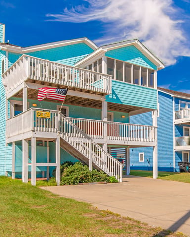 Turquoise beach house with white railings, multiple decks, and American flag under clear blue sky