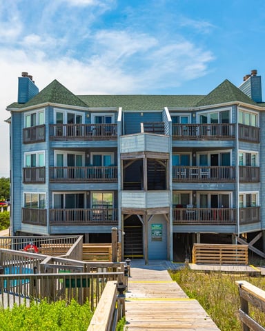 Large blue three-story beach house with green roof, multiple decks, and wooden boardwalk leading to pool area