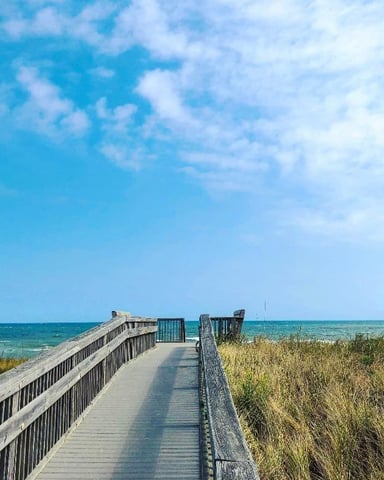 Wooden boardwalk with railings extending toward ocean beach under blue sky with white clouds