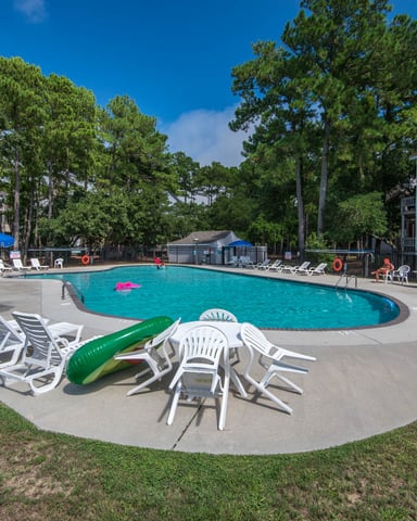 Community pool with turquoise water, white lounge chairs, blue umbrellas, and residential buildings surrounded by tall pine trees