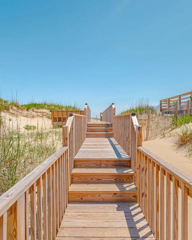 Wooden boardwalk with railings leading through sand dunes with beach grass toward the ocean under clear blue sky