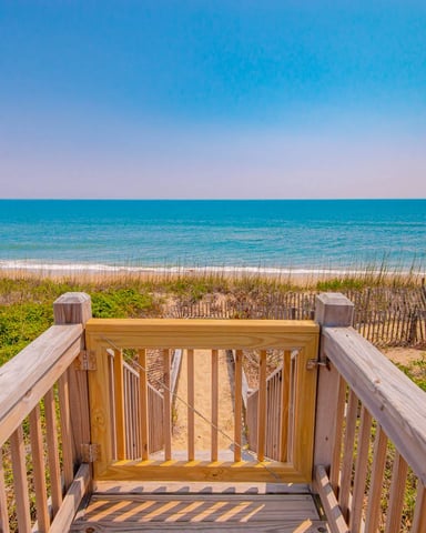 Wooden beach boardwalk with stairs leading down to sandy beach, dune grass, and turquoise ocean under clear blue sky