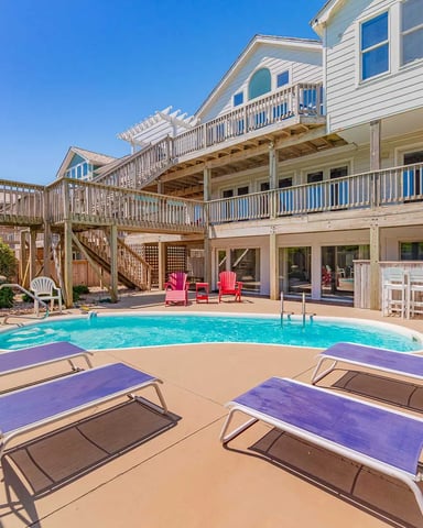 Beachfront house with swimming pool, lounge chairs, multiple decks, and Adirondack seating area under clear blue sky