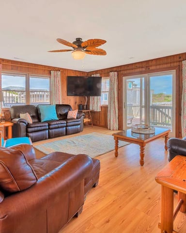 Wood-paneled living room with brown leather seating, ocean views through multiple windows, and coastal decor accents