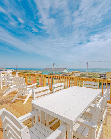 Wooden deck with white Adirondack chairs and dining table overlooking beach and ocean under blue sky