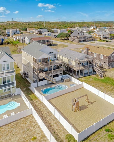 Aerial view of beachfront residential neighborhood with elevated homes, pools, and sandy yards bordered by white fencing