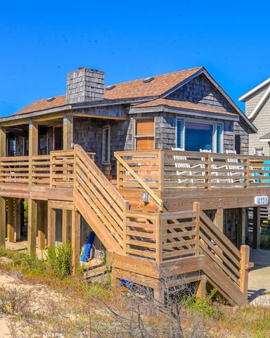 Beachfront cottage with wooden deck and stairs, shingle siding, and neighboring houses under clear blue sky