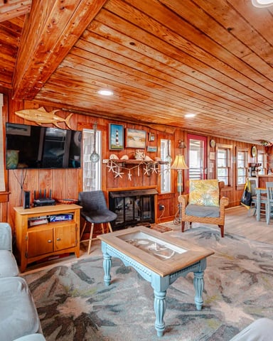 Spacious wooden cabin interior with exposed beam ceiling, nautical decor, fireplace, TV, comfortable seating, and ocean views through windows