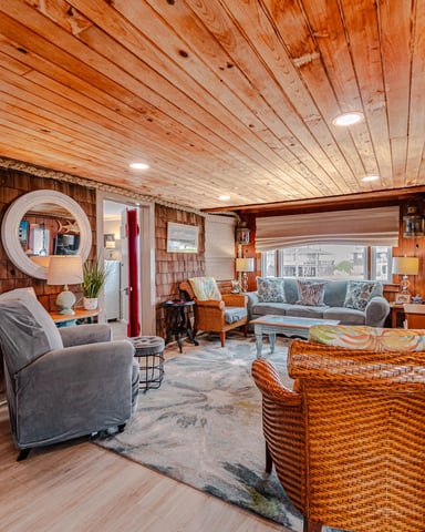 Spacious living room with wood plank ceiling, shingle walls, gray upholstered chairs, woven furniture, and windows overlooking water.