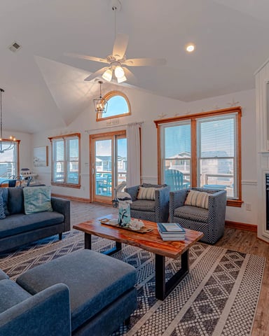 Bright open-plan living room with blue upholstered seating, wood coffee table, fireplace, and multiple windows with ocean views