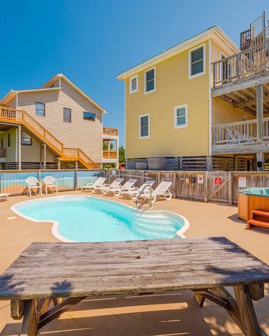 Beachfront vacation rental community with pool, hot tub, lounge chairs, and multi-story beach houses on sandy ground.