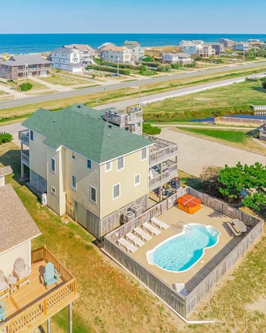Aerial view of yellow beachfront house with green roof, private pool, and wooden deck overlooking ocean and neighboring homes