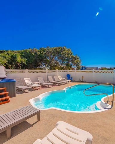 Fenced pool deck with turquoise swimming pool, hot tub, lounge chairs, and multi-story beach house under clear blue sky