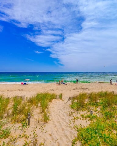 Sandy beach with dune grass, wooden fence, and beachgoers in turquoise ocean under blue sky