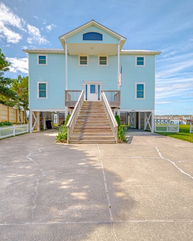 Light blue elevated beach house with wooden deck stairs, white railings, and water view behind white picket fence