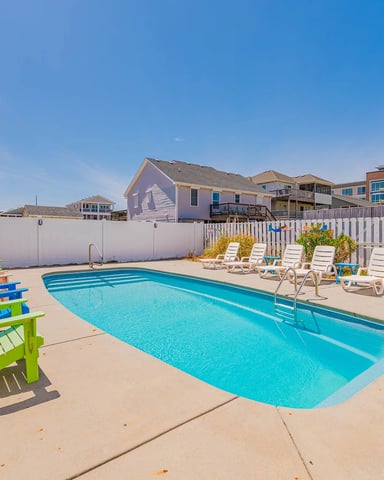 Rectangular swimming pool with bright blue water surrounded by concrete deck, colorful Adirondack chairs, and white fencing, with beachfront houses in