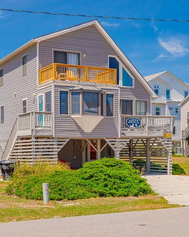 Elevated beach house with gray siding, yellow deck, and wooden pilings in residential neighborhood under clear blue sky