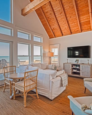 Bright open-plan living room with exposed wood ceiling, ocean view through large windows, beige sofa, wicker chairs, and deck access with Adirondack chairs.