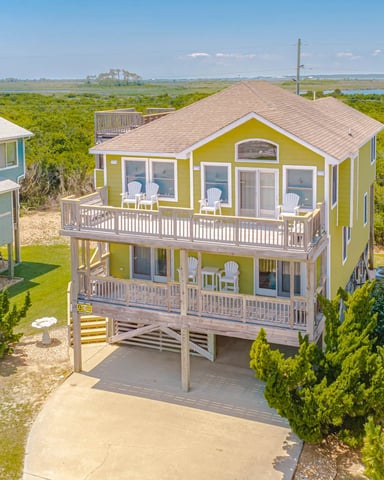 Yellow elevated beach house with wraparound decks overlooking marshland and water, neighboring blue cottage visible left
