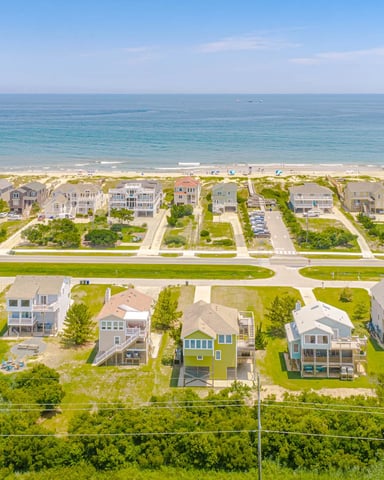 Aerial view of beachfront residential neighborhood with rows of houses, green lawns, sandy beach, and ocean horizon