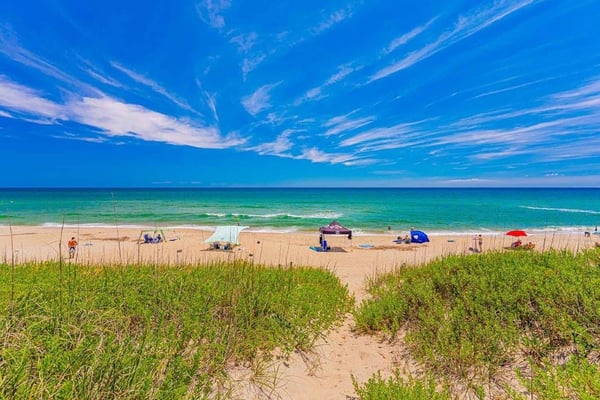 Sandy beach with dune grass, colorful umbrellas, and turquoise ocean under clear blue sky