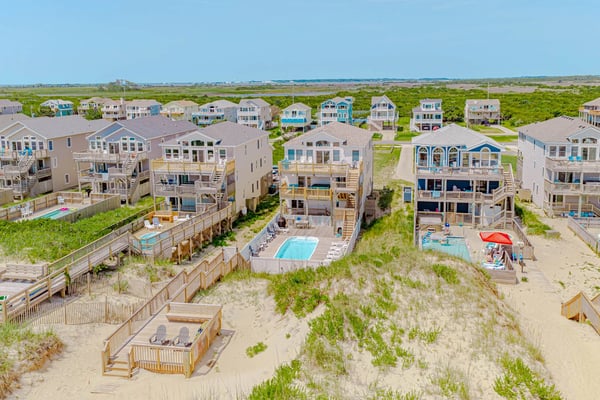 Aerial view of beachfront vacation rental homes with sand dunes, wooden decks, pools, and marshland beyond