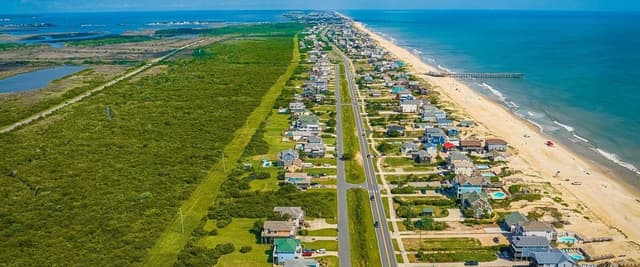 Aerial view of coastal residential community with beach, ocean, and marshland under blue sky with clouds