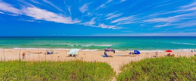 Sandy beach with dune grass, colorful umbrellas and beachgoers, turquoise ocean and blue sky with wispy clouds